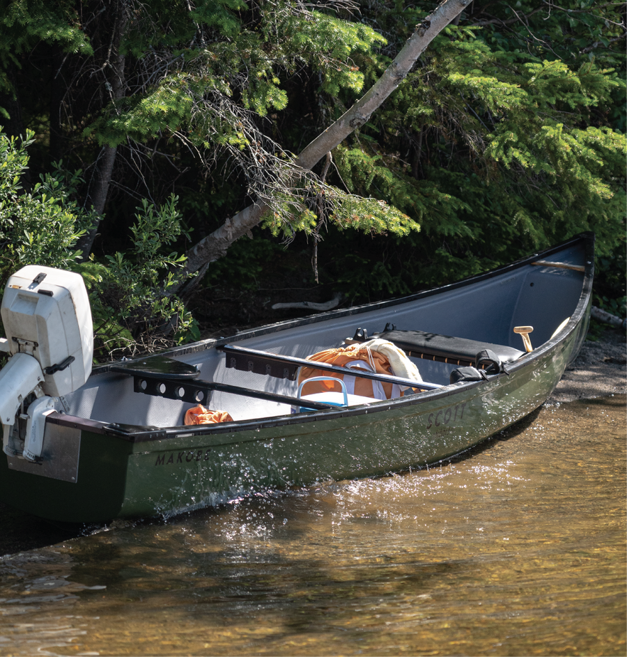 Green Makobe 16' Y-Stern canoe with outboard motor, loaded, at the edge of a forest river